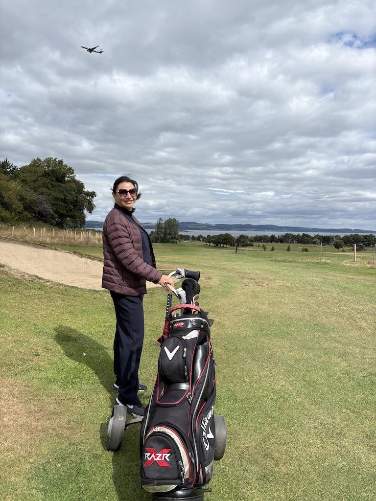 Eteri McKenzie standing on a golf course, smiling confidently beside her golf bag under a wide, open sky.